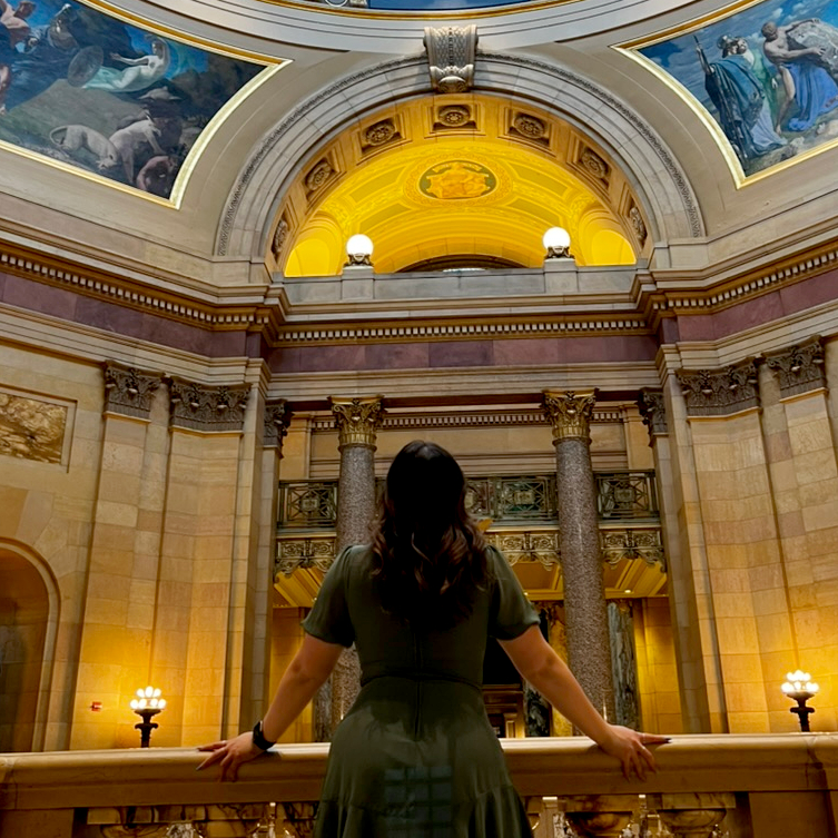 A woman in a green dress, seen from behind, looking up at a gilded rotunda with painted murals