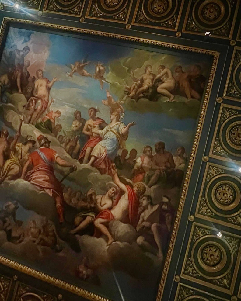 A painted ceiling mural of figures among clouds in a gilded frame, surrounded by ornate coffered panels