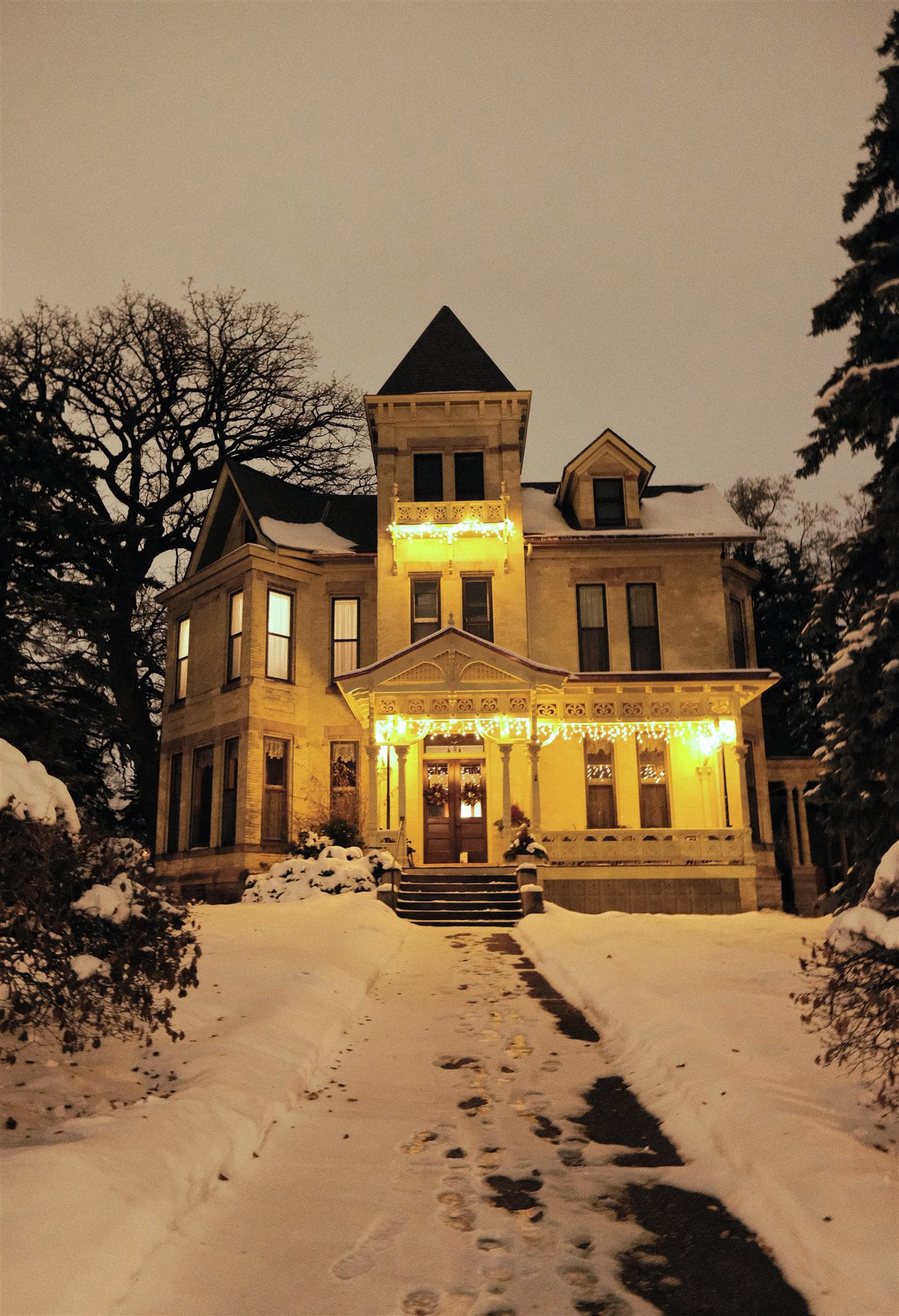 A Victorian house glowing with warm lights at the top of a snowy, footprint-marked path